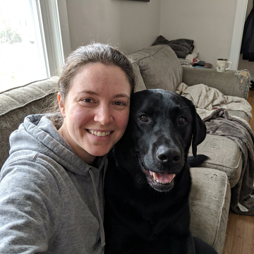 A smiling person hugging a black Labrador on a couch, both faces close